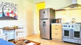 A bright and cozy kitchen featuring a stainless steel refrigerator next to a wooden cabinet with a microwave and a kettle on top. Beneath a wooden shelf lined with glassware, there is a modern gas stove with an overhead exhaust hood. The backsplash consists of white subway tiles. Adjacent to the kitchen is a small dining area with a table set for two, complete with plates and wine glasses. The walls are adorned with a colorful artwork depicting abstract houses under a stylized tree.