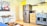 A bright and cozy kitchen featuring a stainless steel refrigerator next to a wooden cabinet with a microwave and a kettle on top. Beneath a wooden shelf lined with glassware, there is a modern gas stove with an overhead exhaust hood. The backsplash consists of white subway tiles. Adjacent to the kitchen is a small dining area with a table set for two, complete with plates and wine glasses. The walls are adorned with a colorful artwork depicting abstract houses under a stylized tree.