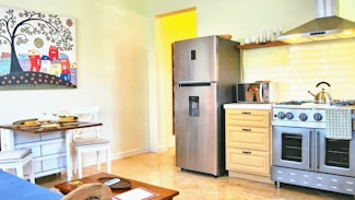 A bright and cozy kitchen featuring a stainless steel refrigerator next to a wooden cabinet with a microwave and a kettle on top. Beneath a wooden shelf lined with glassware, there is a modern gas stove with an overhead exhaust hood. The backsplash consists of white subway tiles. Adjacent to the kitchen is a small dining area with a table set for two, complete with plates and wine glasses. The walls are adorned with a colorful artwork depicting abstract houses under a stylized tree.