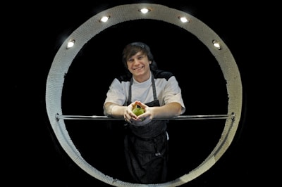 A person with short hair in a chef's uniform stands inside a circular, illuminated apparatus, holding a green apple with a smile. The apparatus is metallic with built-in lights and appears to be part of a kitchen or culinary setting.