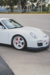 The image features a sleek, white sports car parked on the side of the road. The focus is on the front half of the vehicle, showcasing its aerodynamic design, large alloy wheels, and red brake calipers. In the background, there are trees lining the street, with a blurred view of the pavement and distant fencing.