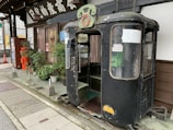 An old, weathered tram or cable car cab is placed on the sidewalk next to a building with traditional signage. There is a variety of green plants in pots in front of the windows and a red vintage post box nearby. The cab has a faded black exterior with rust and chipped paint, and a green sign is mounted on top.