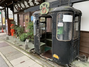 An old, weathered tram or cable car cab is placed on the sidewalk next to a building with traditional signage. There is a variety of green plants in pots in front of the windows and a red vintage post box nearby. The cab has a faded black exterior with rust and chipped paint, and a green sign is mounted on top.