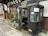 An old, weathered tram or cable car cab is placed on the sidewalk next to a building with traditional signage. There is a variety of green plants in pots in front of the windows and a red vintage post box nearby. The cab has a faded black exterior with rust and chipped paint, and a green sign is mounted on top.