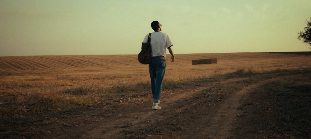 man in white t-shirt and blue denim jeans walking on brown field during daytime