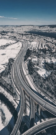 An aerial view of a complex system of highways elegantly weaving through a snow-covered landscape. The roads stretch and curve, guiding a few vehicles amidst the serene winter scenery. A dense urban area and a large body of water are visible in the distance, contrasting with the natural and urban elements.
