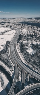 An aerial view of a complex system of highways elegantly weaving through a snow-covered landscape. The roads stretch and curve, guiding a few vehicles amidst the serene winter scenery. A dense urban area and a large body of water are visible in the distance, contrasting with the natural and urban elements.