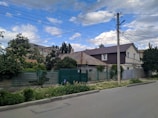 A residential street scene featuring houses with varying architectural styles, surrounded by greenery and fences. Overhead power lines cross above the street, which runs alongside a paved sidewalk. The sky is partly cloudy with patches of blue visible.