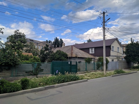 A residential street scene featuring houses with varying architectural styles, surrounded by greenery and fences. Overhead power lines cross above the street, which runs alongside a paved sidewalk. The sky is partly cloudy with patches of blue visible.