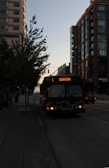 A public bus traveling along a city street with tall buildings and trees lining both sides. The bus displays a destination heading of 'Downtown.' The scene is set during a time of low light, possibly near sunset, giving the environment a dark and calm appearance.