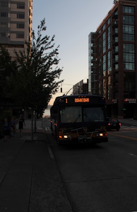 A public bus traveling along a city street with tall buildings and trees lining both sides. The bus displays a destination heading of 'Downtown.' The scene is set during a time of low light, possibly near sunset, giving the environment a dark and calm appearance.