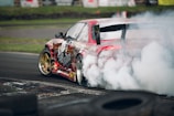 A red sports car is drifting on a racetrack, producing a large cloud of smoke from its rear tires. The car features distinctive graphics and gold rims, and it speeds around a corner surrounded by worn tires. The background includes a grassy area and blurred signage.