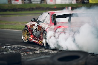 A red sports car is drifting on a racetrack, producing a large cloud of smoke from its rear tires. The car features distinctive graphics and gold rims, and it speeds around a corner surrounded by worn tires. The background includes a grassy area and blurred signage.