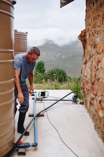 A man is working near large water tanks connected by pipes. He is wearing a gray shirt and dark pants, and the setting is outdoors with a mountainous landscape in the background. The ground is concrete, and there is a stone wall on the right side.
