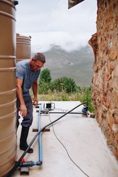 A man is working near large water tanks connected by pipes. He is wearing a gray shirt and dark pants, and the setting is outdoors with a mountainous landscape in the background. The ground is concrete, and there is a stone wall on the right side.