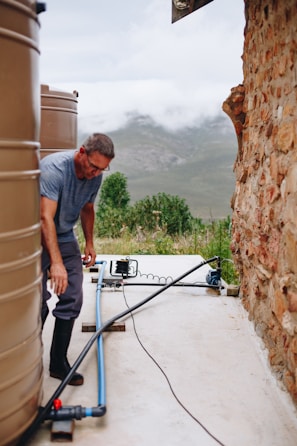 A man is working near large water tanks connected by pipes. He is wearing a gray shirt and dark pants, and the setting is outdoors with a mountainous landscape in the background. The ground is concrete, and there is a stone wall on the right side.