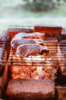 Several raw meat steaks are positioned on a metal grill over glowing coals within a rustic outdoor barbecue setup. Surrounding the grill are bricks forming a makeshift barbecue pit.