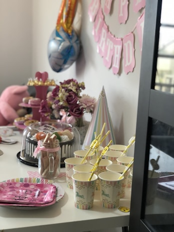 A festive birthday setup with pink and pastel decorations. Balloons are seen in the background alongside a pink 'Happy Birthday' banner on the wall. A table is adorned with various elements, including a tiered cupcake stand, a cake under a glass dome, plates, cups with yellow straws, and a party hat. Floral arrangements add a touch of elegance to the decor.