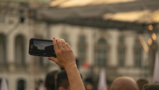 A person holding a smartphone aloft, seemingly capturing a photo or video. The background shows an out-of-focus building with multiple windows and arched doorways, and there seems to be a gathering or event taking place.