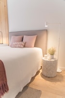 A cozy bedroom setting features a neatly made bed with a light gray upholstered headboard, decorated with soft pink pillows and a decorative cushion. Beside the bed, a round woven nightstand holds a white candle and a vase with dried plants, illuminated by a white standing lamp. The wall behind is accented with vertical wooden slats, while the floor showcases light wood with a small woven rug.