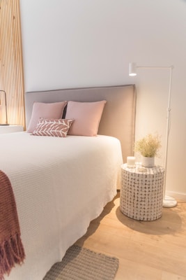 A cozy bedroom setting features a neatly made bed with a light gray upholstered headboard, decorated with soft pink pillows and a decorative cushion. Beside the bed, a round woven nightstand holds a white candle and a vase with dried plants, illuminated by a white standing lamp. The wall behind is accented with vertical wooden slats, while the floor showcases light wood with a small woven rug.