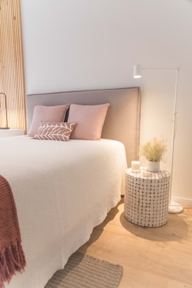 A cozy bedroom setting features a neatly made bed with a light gray upholstered headboard, decorated with soft pink pillows and a decorative cushion. Beside the bed, a round woven nightstand holds a white candle and a vase with dried plants, illuminated by a white standing lamp. The wall behind is accented with vertical wooden slats, while the floor showcases light wood with a small woven rug.