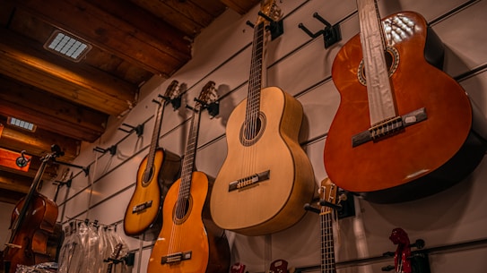Several classical guitars hang on a display wall, with acoustic bodies made from light and dark wood. The setting appears to be a music store with a wooden ceiling and other musical instruments visible on the sides.