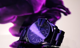 A sleek, metallic wristwatch with a dark dial is prominently displayed against a soft, abstract purple background. The watch has a stainless steel band and glowing hour markers, with light reflecting off its surface, adding a sense of luxury.