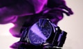 A sleek, metallic wristwatch with a dark dial is prominently displayed against a soft, abstract purple background. The watch has a stainless steel band and glowing hour markers, with light reflecting off its surface, adding a sense of luxury.