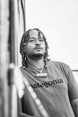 A person with braided hair and a thoughtful expression, wearing a t-shirt that reads 'Patagonia' and a necklace, is leaning against a textured wall. The image is in black and white, creating a contrast between the subject and the background.