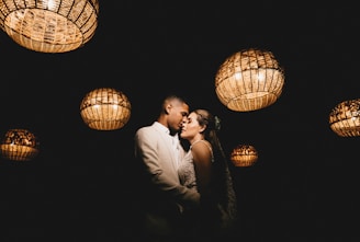 A couple is embracing intimately under a series of hanging, woven basket-style lights that illuminate them softly against a dark background. The scene has a romantic and intimate ambiance.