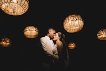 A couple is embracing intimately under a series of hanging, woven basket-style lights that illuminate them softly against a dark background. The scene has a romantic and intimate ambiance.