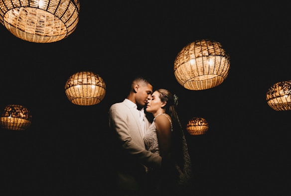 A couple is embracing intimately under a series of hanging, woven basket-style lights that illuminate them softly against a dark background. The scene has a romantic and intimate ambiance.