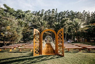 An outdoor wedding setting is arranged with a rustic wooden archway and benches on a grassy lawn surrounded by lush trees. The clear sky and sunlight create a peaceful and inviting atmosphere, with the aisle lined with flowers.