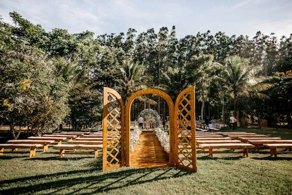 An outdoor wedding setting is arranged with a rustic wooden archway and benches on a grassy lawn surrounded by lush trees. The clear sky and sunlight create a peaceful and inviting atmosphere, with the aisle lined with flowers.