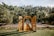 An outdoor wedding setting is arranged with a rustic wooden archway and benches on a grassy lawn surrounded by lush trees. The clear sky and sunlight create a peaceful and inviting atmosphere, with the aisle lined with flowers.