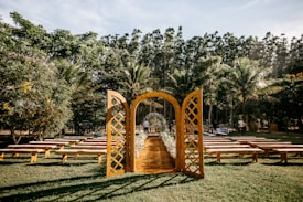 An outdoor wedding setting is arranged with a rustic wooden archway and benches on a grassy lawn surrounded by lush trees. The clear sky and sunlight create a peaceful and inviting atmosphere, with the aisle lined with flowers.