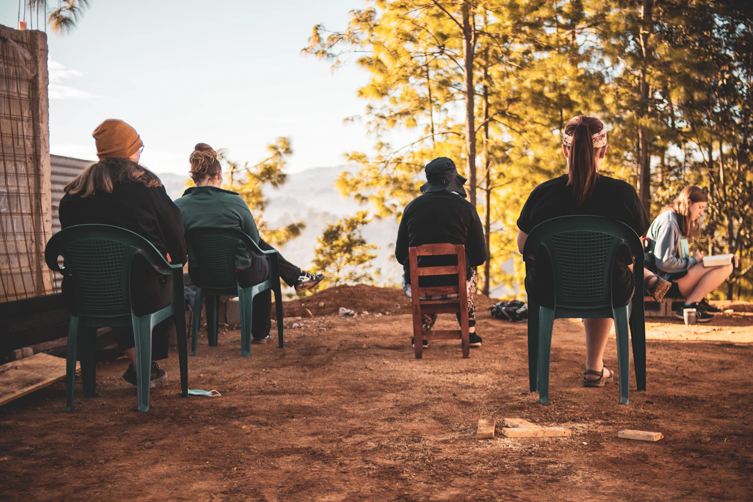 People sitting on plastic chairs on bare ground in low gold light, backs to the camera, facing a speaker out of frame