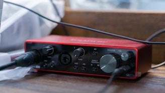 A compact audio interface with a red casing is situated on a wooden surface. It has various knobs and ports for gain, monitor control, and direct input, with cables plugged into some of the ports. The lighting creates a soft, focused effect on the device, with a blurred background.