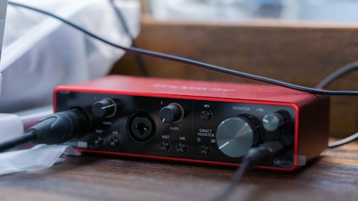 A compact audio interface with a red casing is situated on a wooden surface. It has various knobs and ports for gain, monitor control, and direct input, with cables plugged into some of the ports. The lighting creates a soft, focused effect on the device, with a blurred background.