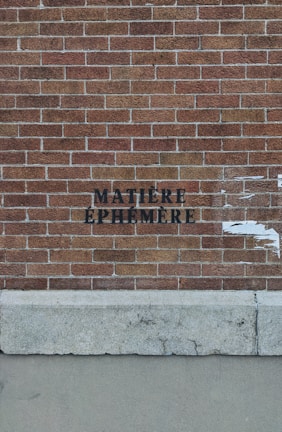 A red brick wall with the words 'MATIÈRE ÉPHÉMÈRE' stenciled in black paint. The lower section of the image shows a concrete base with some weathered texture.
