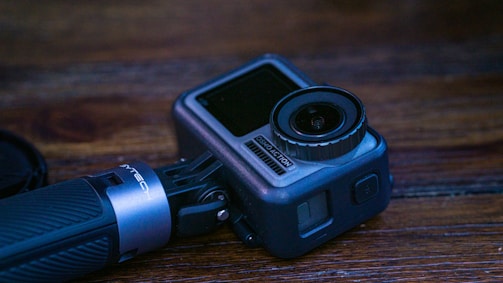 A compact action camera with a textured grip is placed on a wooden surface. The camera has a prominent lens and a side screen, typical of portable filming devices.