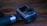 A compact action camera with a textured grip is placed on a wooden surface. The camera has a prominent lens and a side screen, typical of portable filming devices.