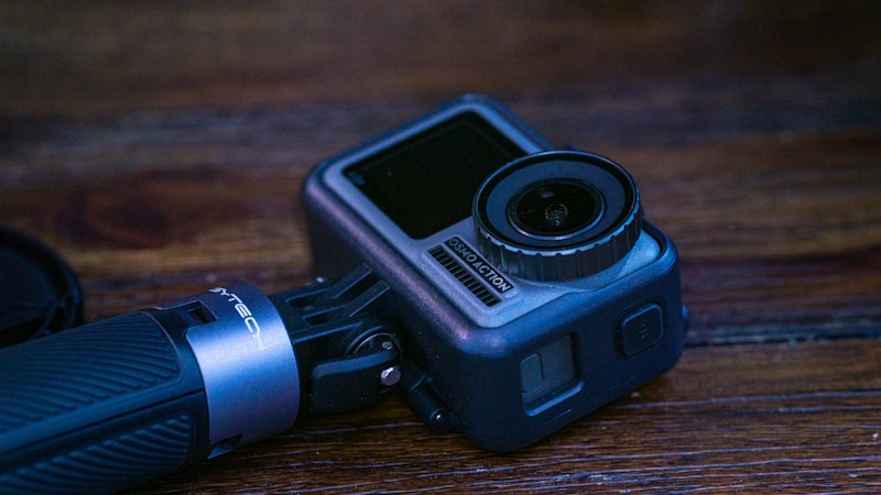 A compact action camera with a textured grip is placed on a wooden surface. The camera has a prominent lens and a side screen, typical of portable filming devices.