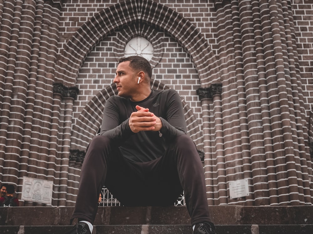 man in black long sleeve shirt and black pants sitting on brown brick wall