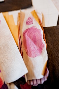 A collection of textured materials and paper featuring a prominent pink brushstroke on one piece. The materials exhibit an array of colors, primarily pinks and browns, with some pieces displaying textured patterns.