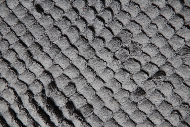 A close-up view of a textured surface featuring a pattern of interwoven, rounded fibers that create a quilted or braided appearance. The surface appears soft and slightly fuzzy, emphasizing the tactile quality of the material.