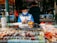 A vendor wearing a blue and white jacket and a face mask is sitting behind a stall filled with various colorful carved seals and objects. The setting appears to be an outdoor market or shop with other items and a person in a dark coat seen in the background. There are papers and a QR code on the countertop, and several red decoration elements are visible.