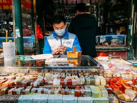 A vendor wearing a blue and white jacket and a face mask is sitting behind a stall filled with various colorful carved seals and objects. The setting appears to be an outdoor market or shop with other items and a person in a dark coat seen in the background. There are papers and a QR code on the countertop, and several red decoration elements are visible.