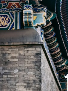 Detailed architectural detail of a traditional structure featuring ornate and colorful decorations on the roofline. The design includes intricate patterns, rich colors, and traditional motifs, with a sturdy grey brick wall beneath.